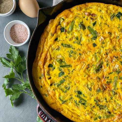 close up of a vegetable frittata in a cast iron pan with colorful hen's eggs to one side and a dish of salt and pepper