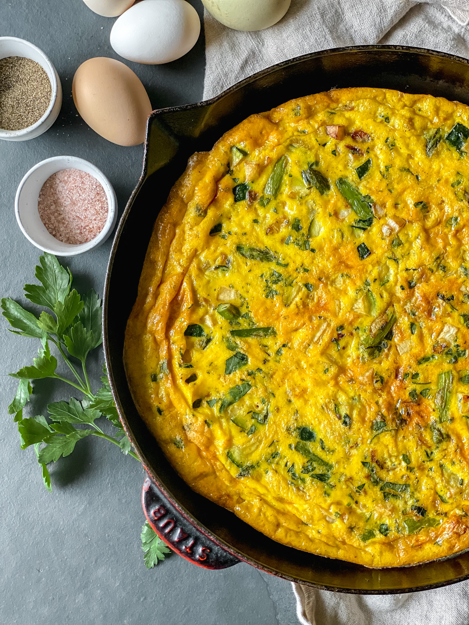 close up of a vegetable frittata in a cast iron pan with colorful hen's eggs to one side and a dish of salt and pepper