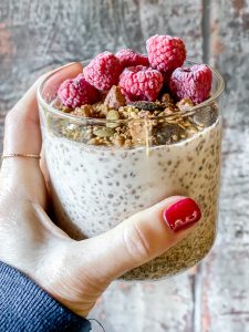 hand with red nails holding glass jar of no oats topped with granola and raspberries