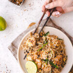 plate of pad thai with lime wedge and hands holding chopsticks